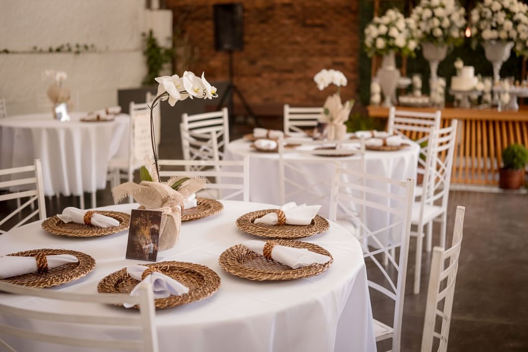Mesas redondas com toalha branca, sousplats de palha e centro de orquídeas brancas